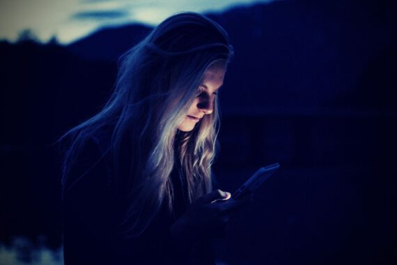 Das Bild zeigt eine blonde Frau, wie sie im Freien auf ihr Handy blickt. Das Foto illustriert einen Artikel auf Deutsch zum Thema Doomscrolling und wie man damit aufhören kann.